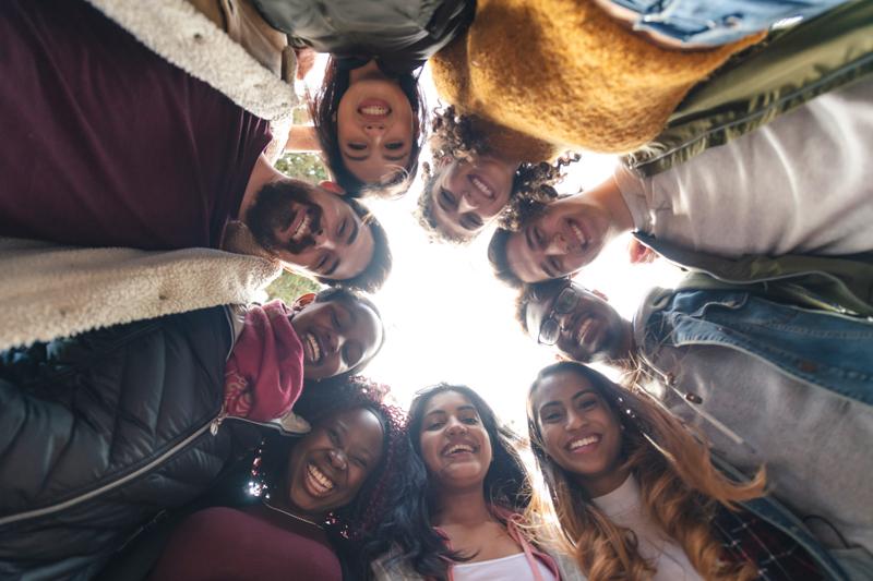 Image of a diverse group of college aged students standing in a circle looking down and smiling at a camera in between them all.
